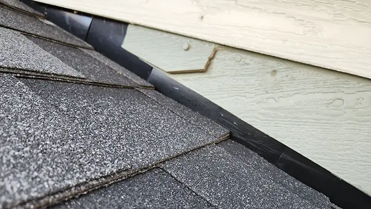 Step flashing 2 - Custom Exteriors, LLC Close-up of dark gray asphalt roof shingles meeting a light-colored wood siding at the roof edge.